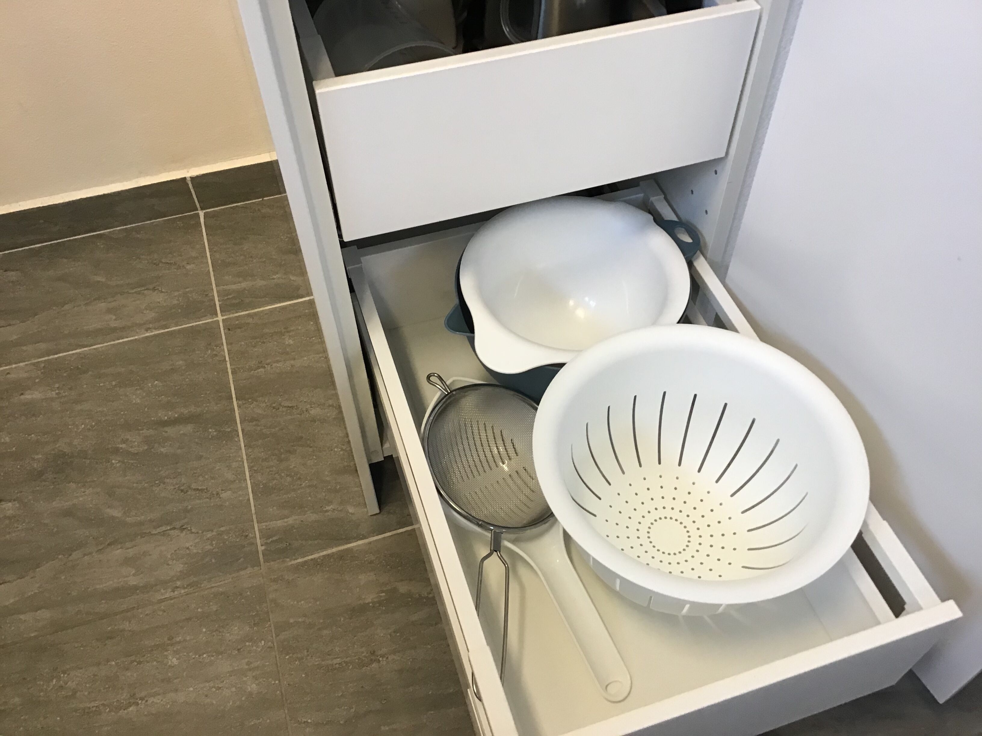 En kökslåda som innehåller vita köksredskap, bland annat ett durkslag, en sil och en skål.