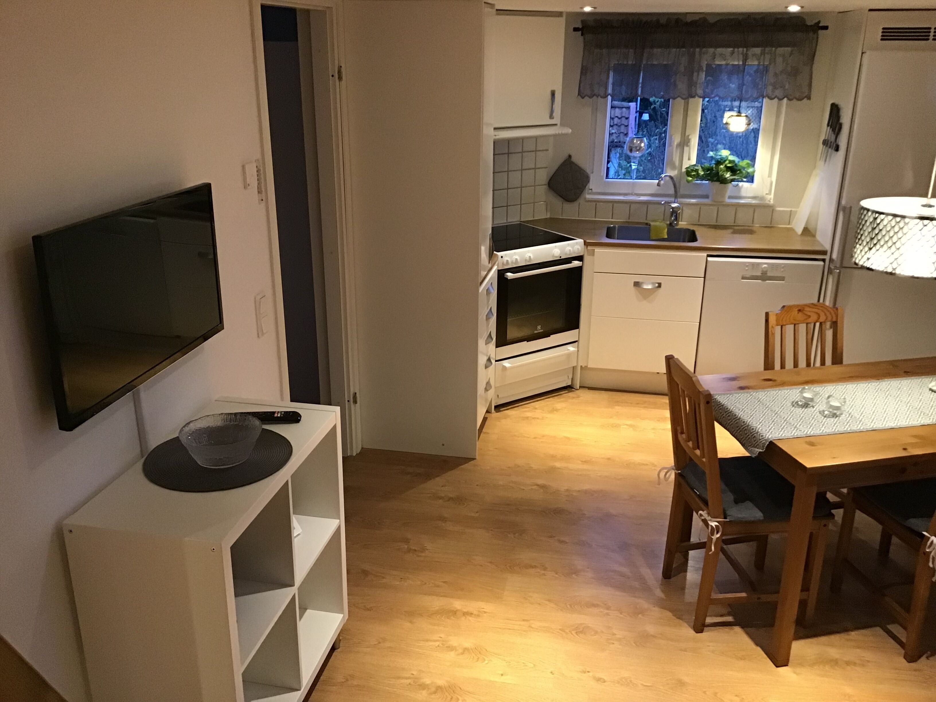 A compact kitchen with a built-in oven, a dining table with chairs, and a TV mounted on the wall.
