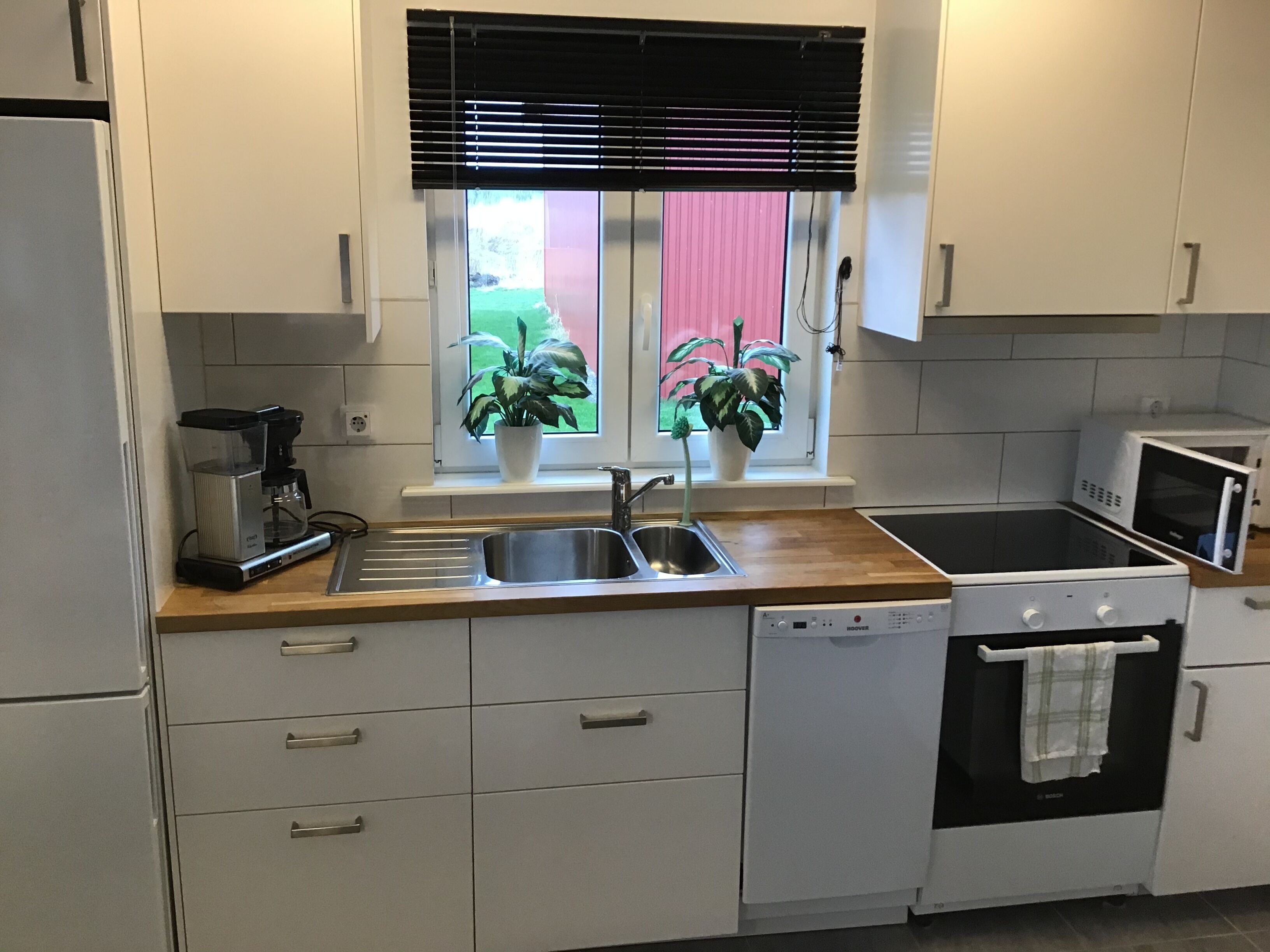 Una cocina moderna con armarios blancos, un lavamanos de acero inoxidable, lavavajillas, microondas y una ventana con persianas.
