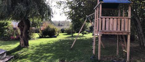 A wooden play structure with a blue roof in a garden with a large tree and grass.