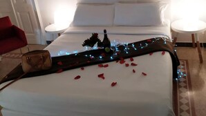 Una cama bien tendida con sábanas blancas, una botella decorativa y pétalos de rosa sobre un suelo alfombrado.