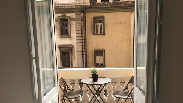 Una puerta abierta que da a un balcón con una mesa redonda y cuatro sillas, una planta en maceta y un edificio con arquitectura clásica al fondo.