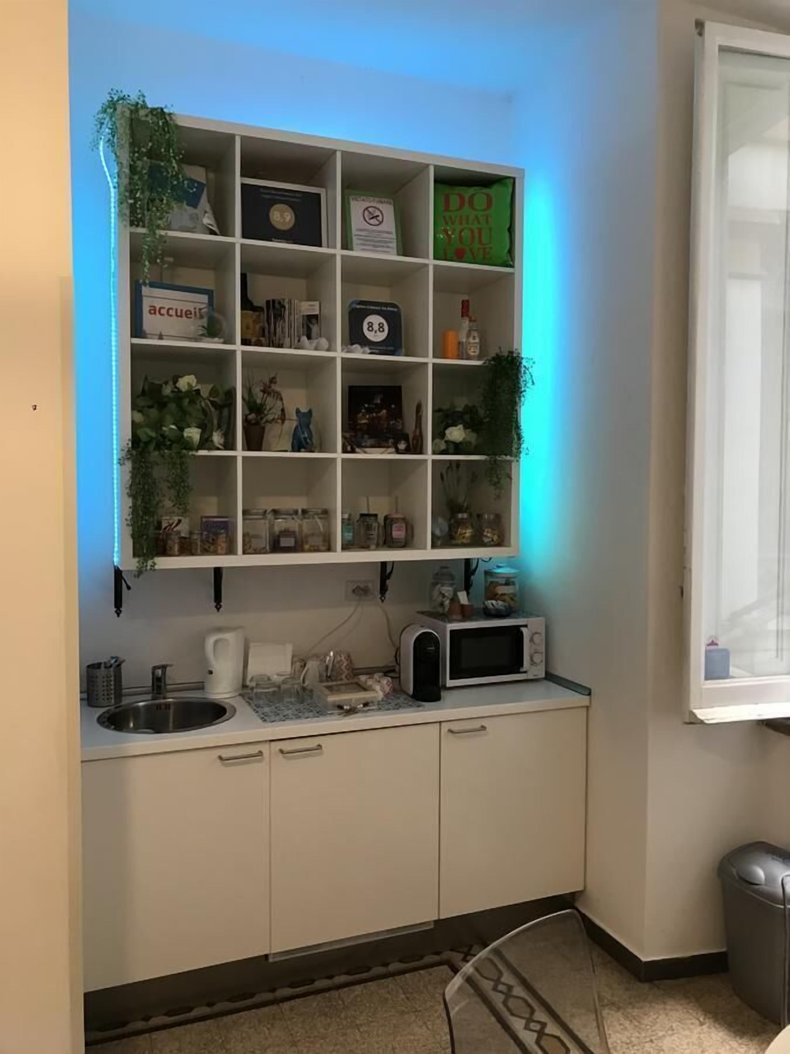 Eine kompakte Küchenzeile mit weißen Schränken, einer Mikrowelle, einem Spülbecken und einer kleinen Arbeitsfläche. Über dem Spülbecken befinden sich Regale mit verschiedenen Gegenständen, darunter eine grüne Tasche und ein gerahmtes Bild.
