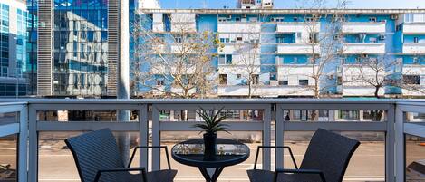 Un balcón con dos sillas y una mesa, con vista a una calle con edificios y árboles.