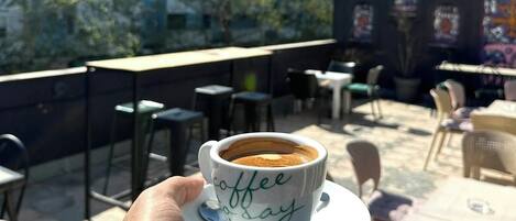 Een hand die een koffiekop vasthoudt, met daarop de tekst 'coffee day', voor een achtergrond van terrasjes en gebouwen.