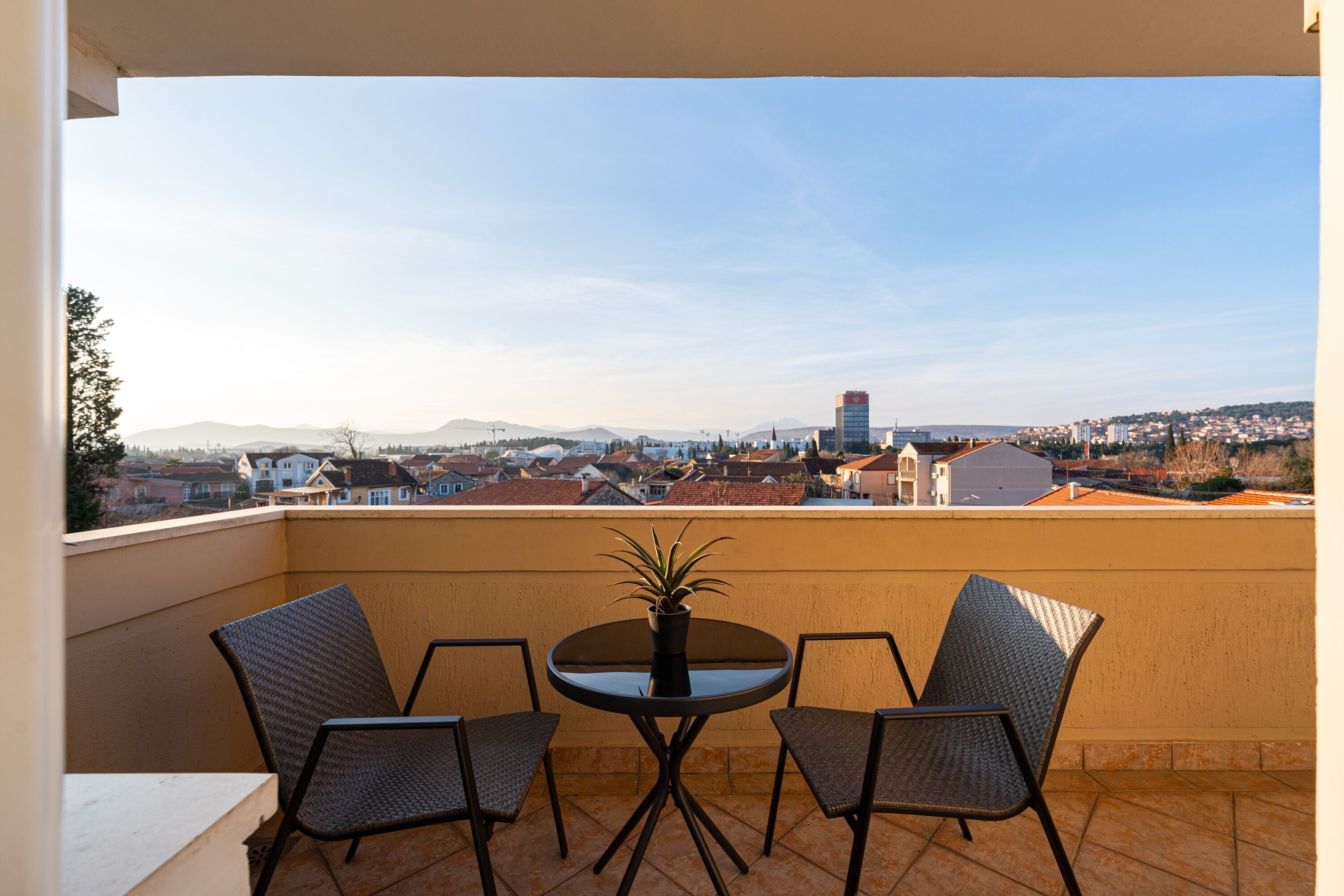 Un balcone con due sedie e un tavolo, affaccia su un panorama urbano con edifici e montagne.