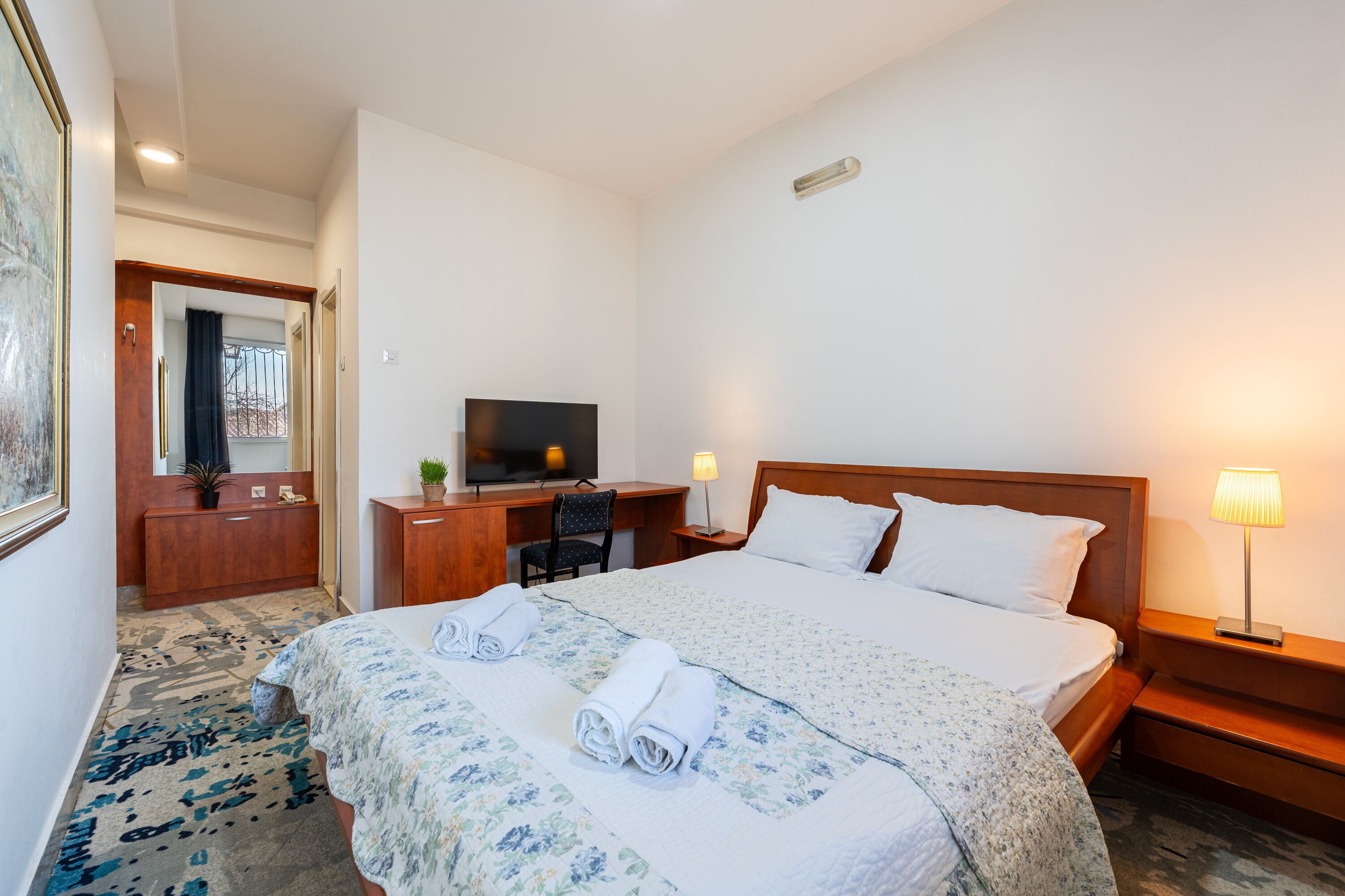 Habitación de hotel con cama, mesita de noche, lámpara, escritorio con televisor y espejo.