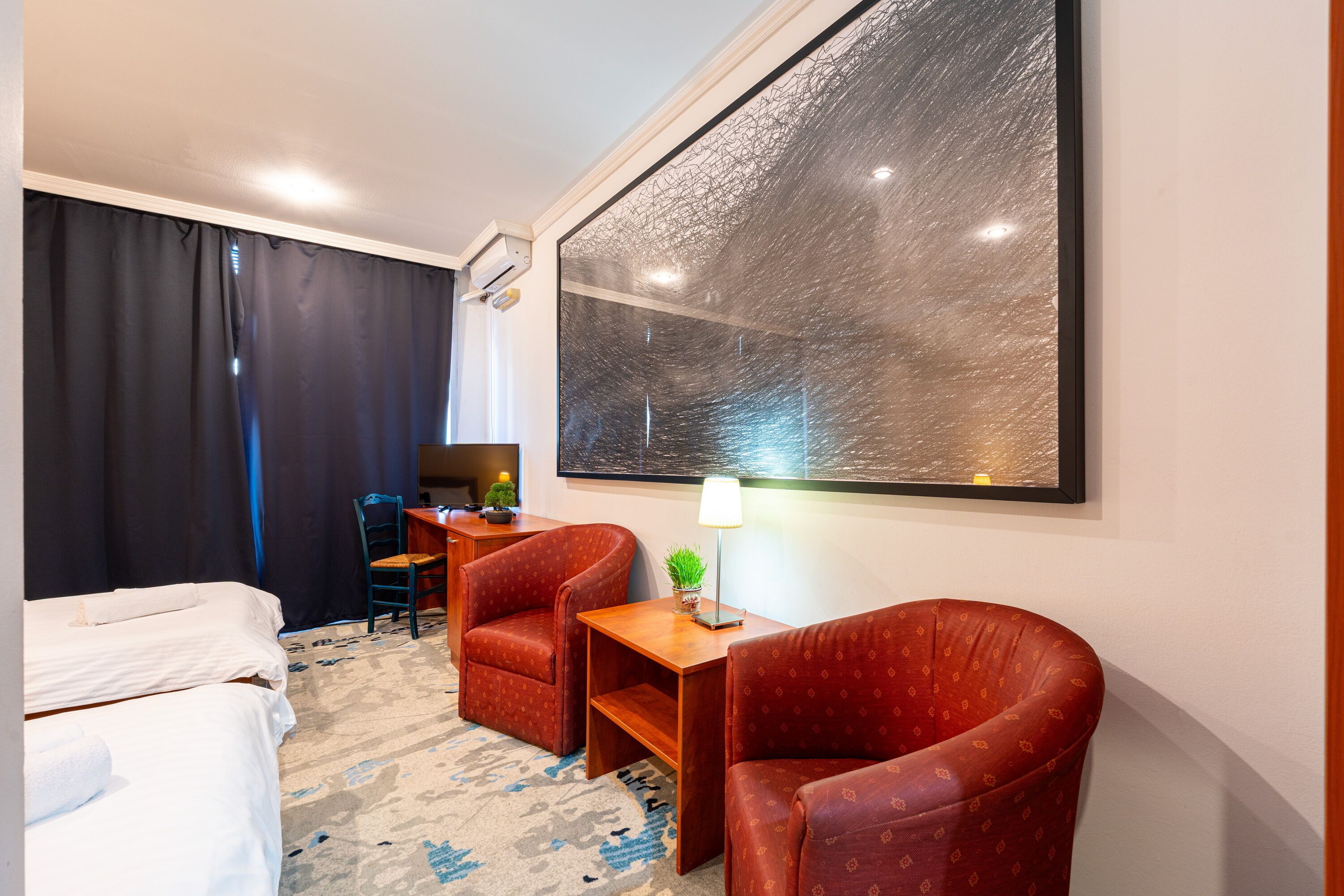 A hotel room with a bed, two armchairs, a small table with a lamp, a desk with a computer, and a large abstract painting on the wall.