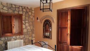 A bedroom with a stone wall, a bed with a white bedspread, a wooden wardrobe, and a mirror with an arch design. - Casita Vista Alegre. (castril)