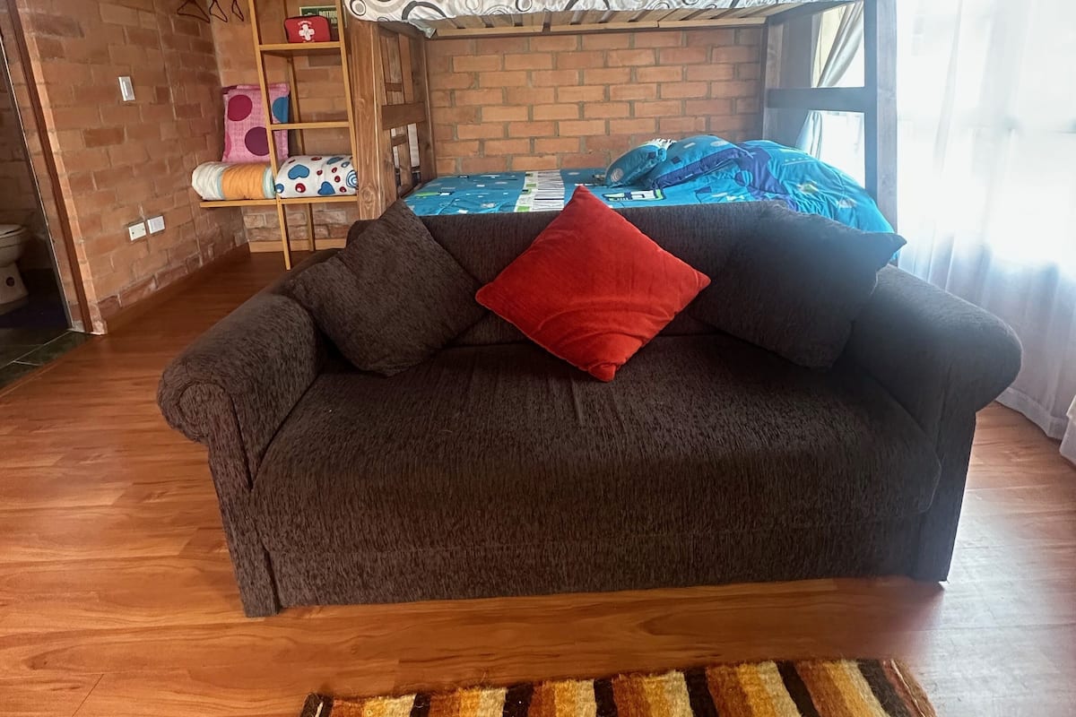 A cozy room with a bunk bed, a sofa with a red pillow, and a wooden floor.