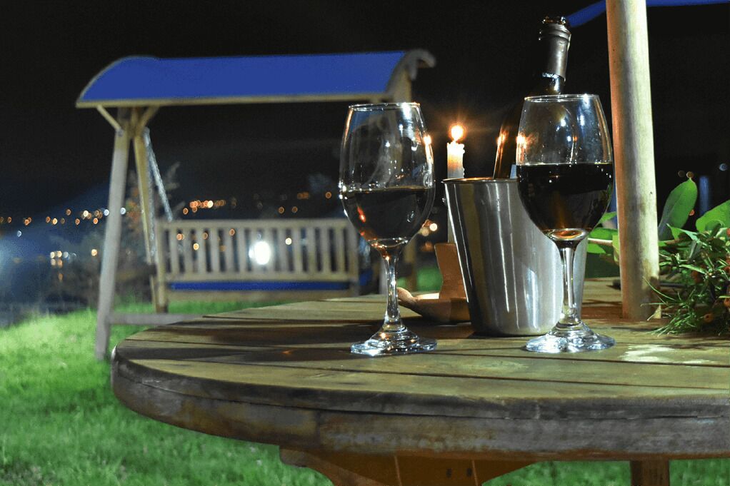A wooden table with two wine glasses and a bottle, a candle, and a metal container, set outdoors at night.