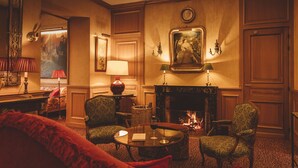 Une pièce chaleureuse, éclairée d’une lumière douce, avec une cheminée, deux fauteuils, un canapé rouge et une table sur laquelle se trouve un verre de vin.