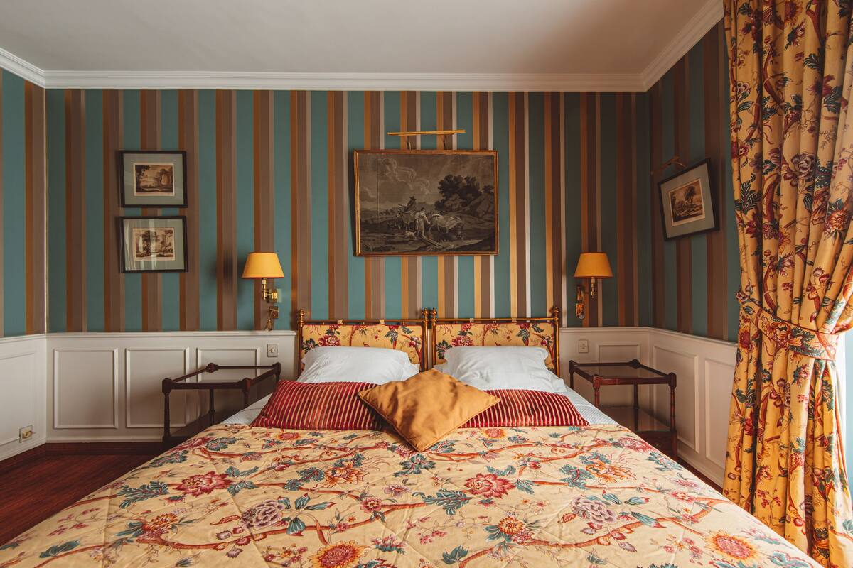 Een bed met een bloemenprint sprei, gestreepte kussens en een houten hoofdbord. Er zijn twee nachtkastjes met lampen.