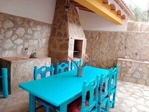 Een overdekte eethoek in openlucht met stenen muren, een blauwe tafel en stoelen, een stenen oven en een gele balkenconstructie.
