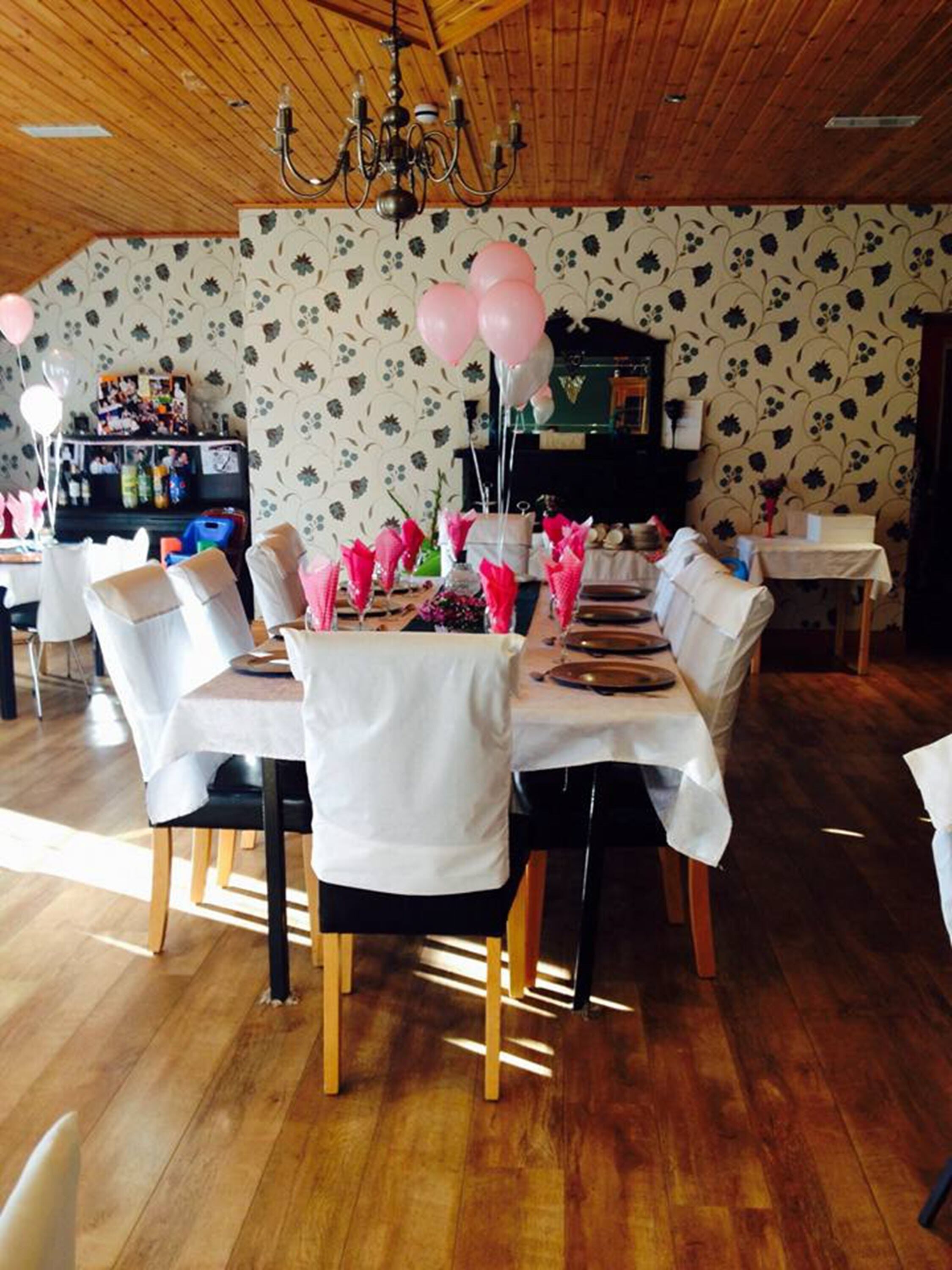 A dining area with a wooden ceiling, floral wallpaper, and a chandelier. Tables are set with white tablecloths and pink napkins.