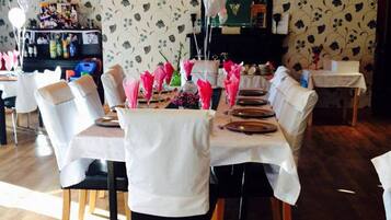 A dining area with a wooden ceiling, floral wallpaper, and a chandelier. Tables are set with white tablecloths and pink napkins.