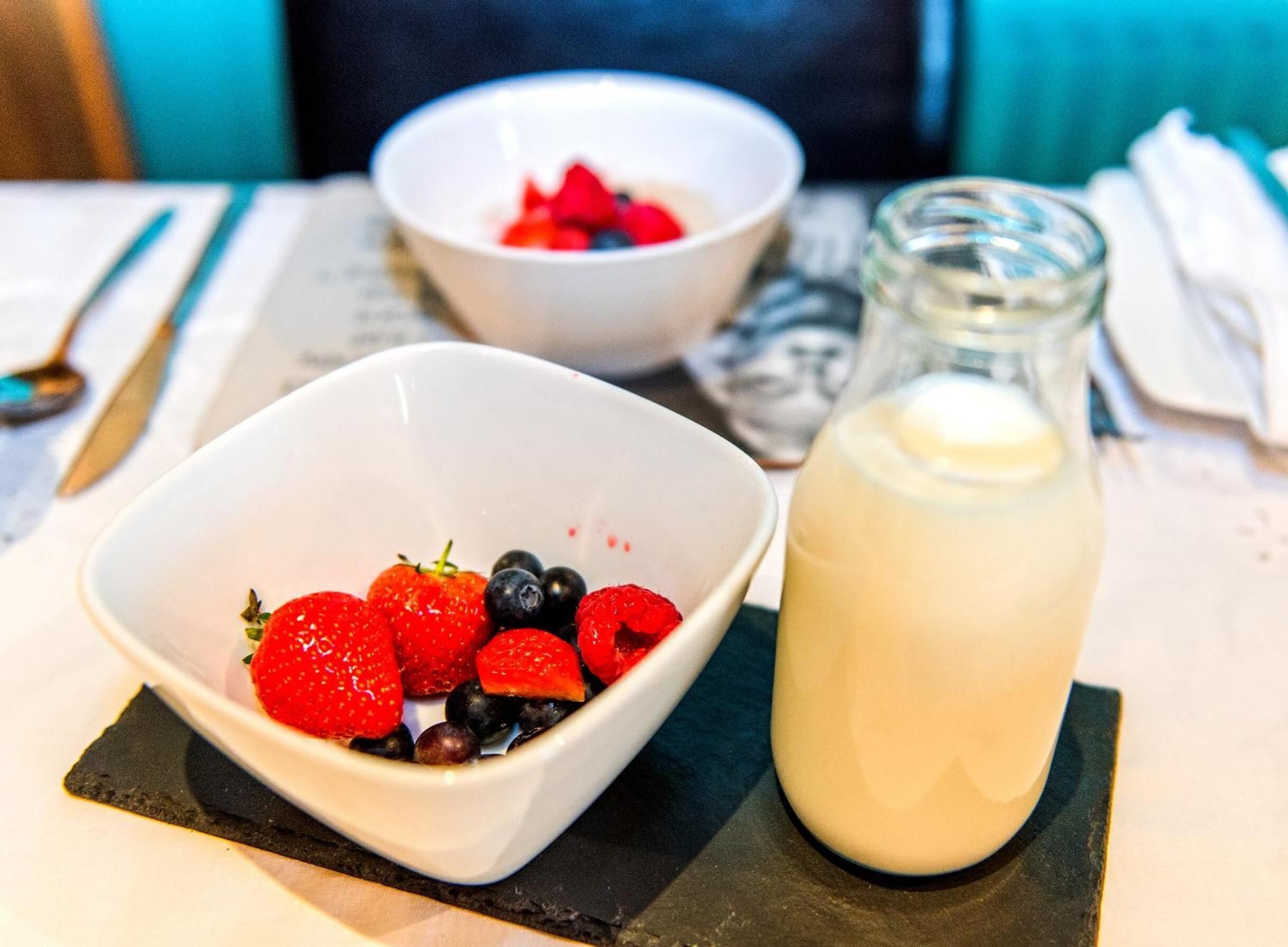 Two white bowls on a table, one with strawberries and blueberries, and a glass bottle of milk.