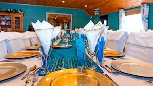 A long dining table set with gold plates, blue napkins, and clear glassware, in a room with wooden ceiling and teal walls.
