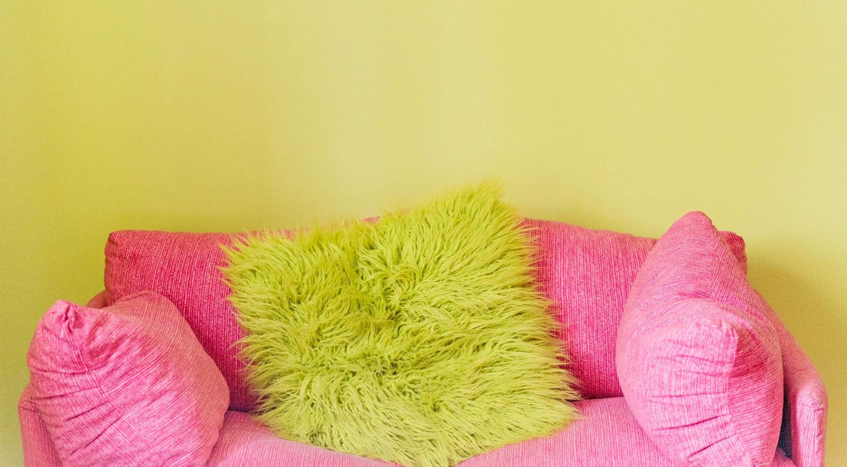 A chair with a fluffy green pillow and two pink pillows against a yellow wall.