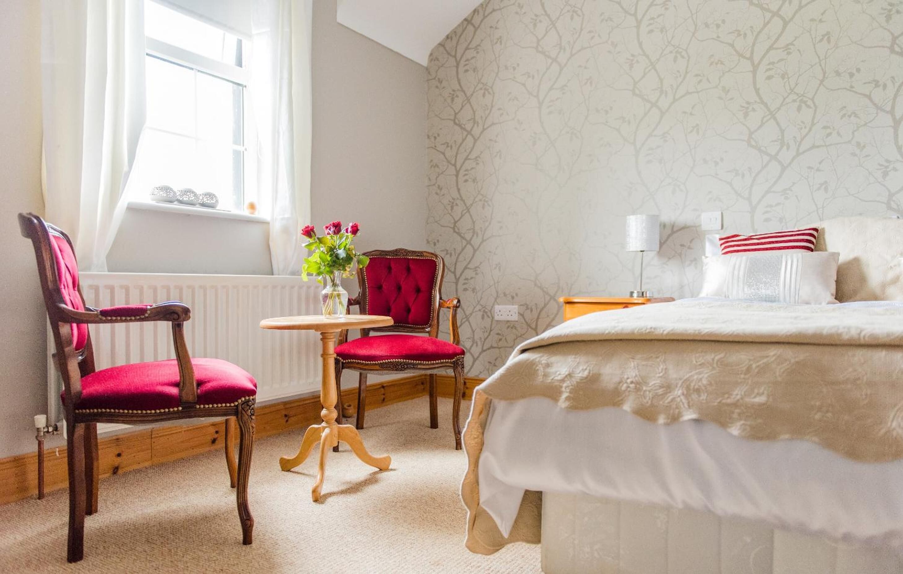 A bedroom with a large bed, two chairs, a small table, and a window with curtains.