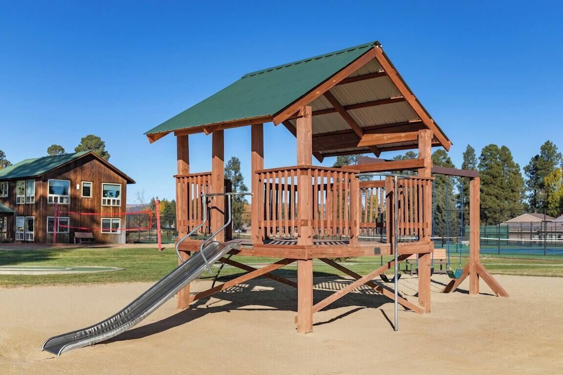Une structure de jeux en bois, dotée d’un toboggan et d’un abri, installée dans un parc avec un bâtiment en arrière-plan.
