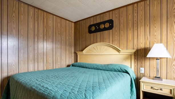 A room with wooden panel walls, a bed with a quilted teal bedspread, a nightstand with a lamp, and a wall-mounted clock. - OYO Hotel Holly Springs MS (Holly Springs)