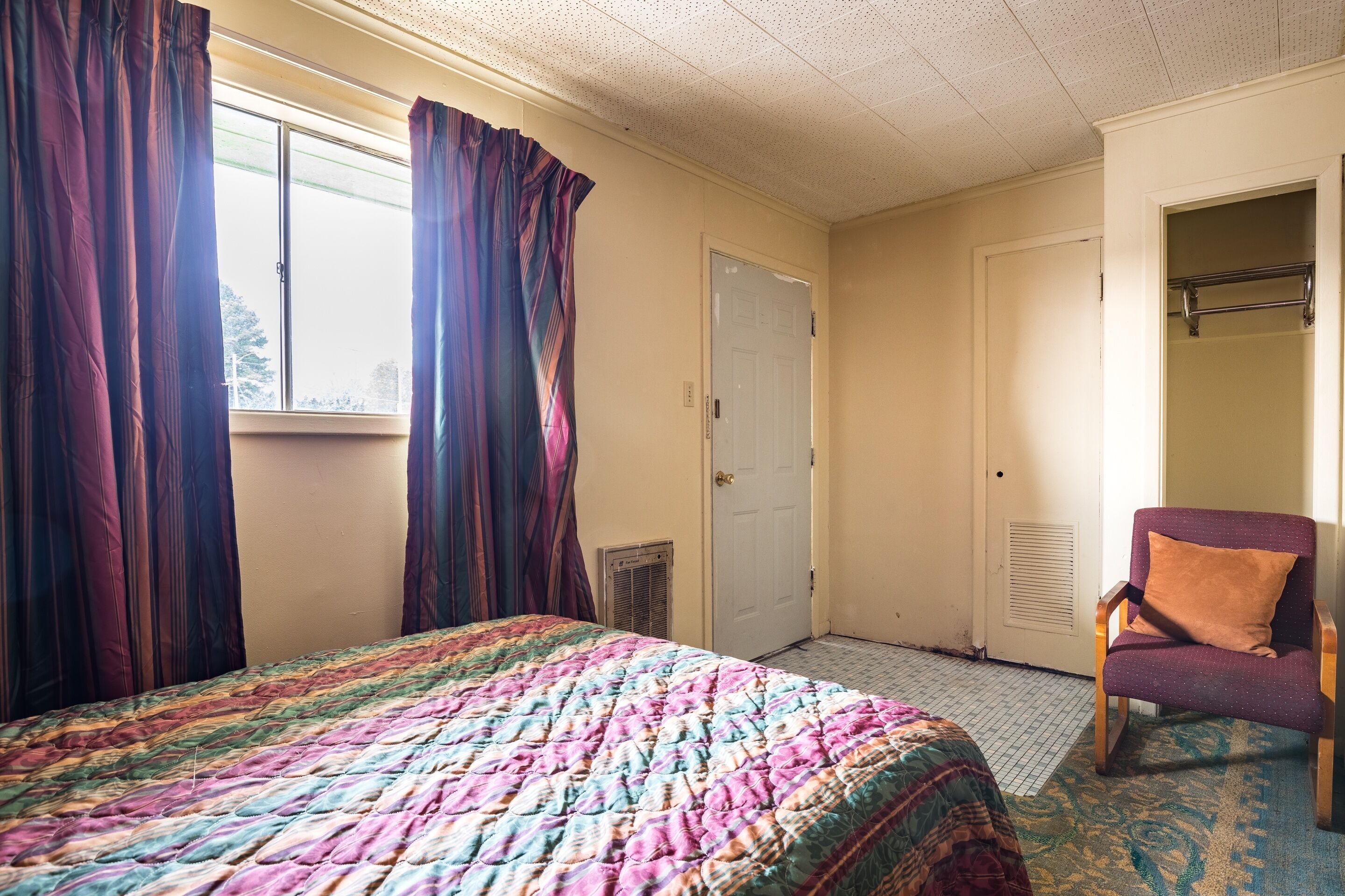 A bedroom with a bed, a chair, a window with curtains, and a door.