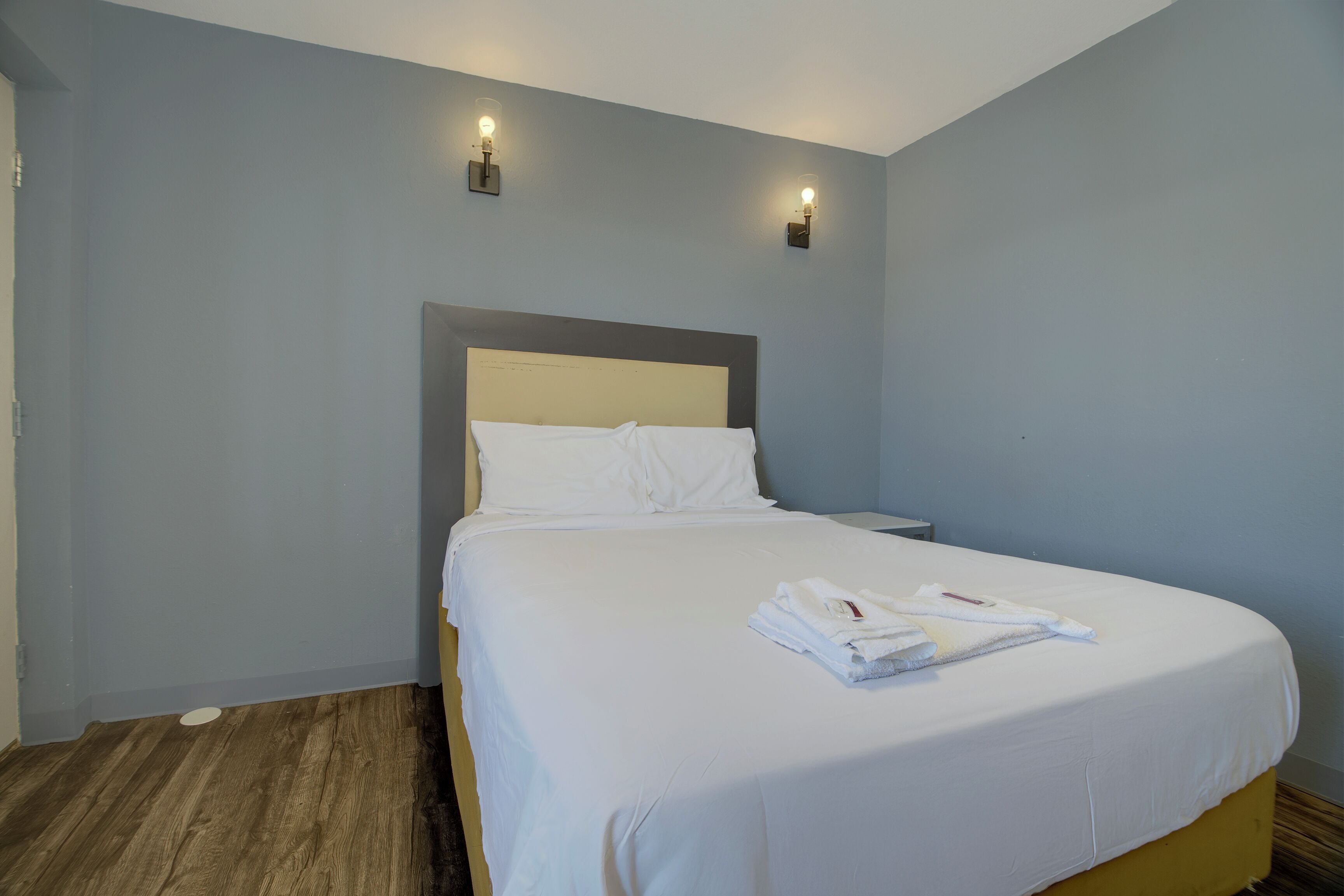 A neatly made bed with white linens and a folded towel, a wooden floor, and light blue walls with two wall-mounted light fixtures.