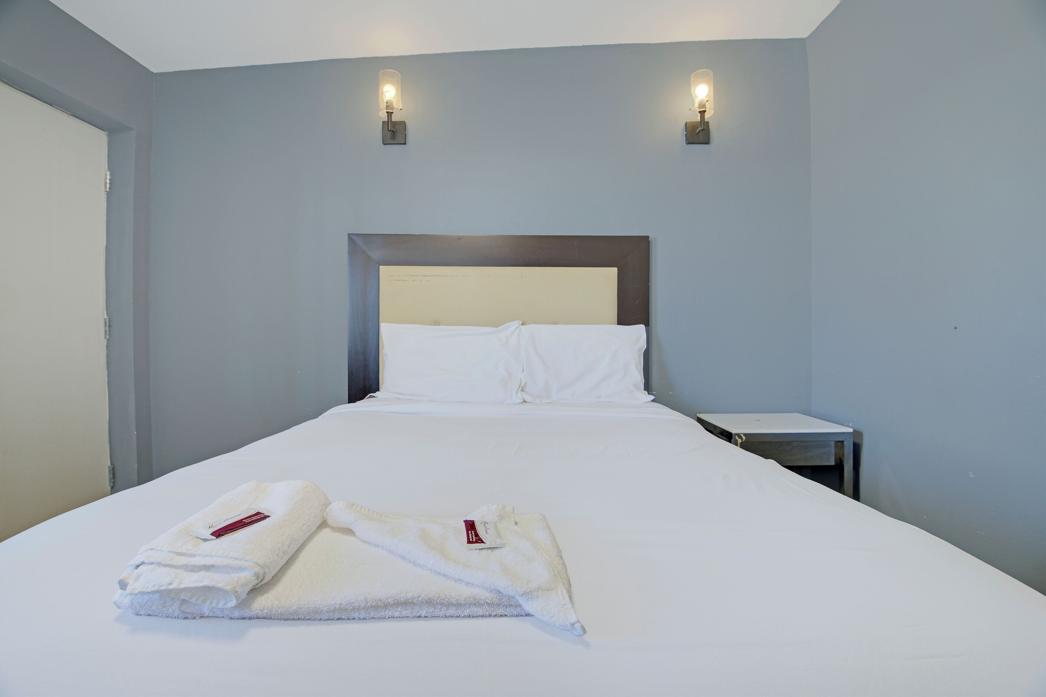 Habitación de hotel con una cama individual, dos lámparas de noche, una mesita de noche y una toalla blanca doblada con cuidado sobre la cama.