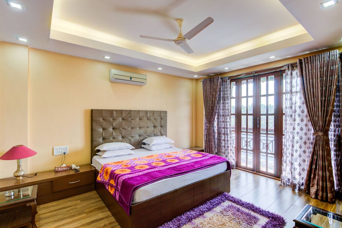 Une chambre à coucher comprenant un lit, un ventilateur de plafond, un appareil de climatisation, une table de chevet, une lampe et une fenêtre avec des rideaux.
