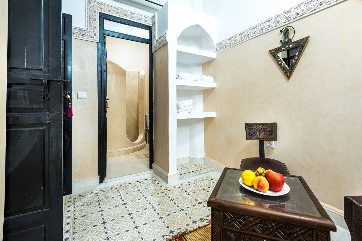 A room with a black door, a shower, a white shelf, a decorative mirror, and a table with fruit.