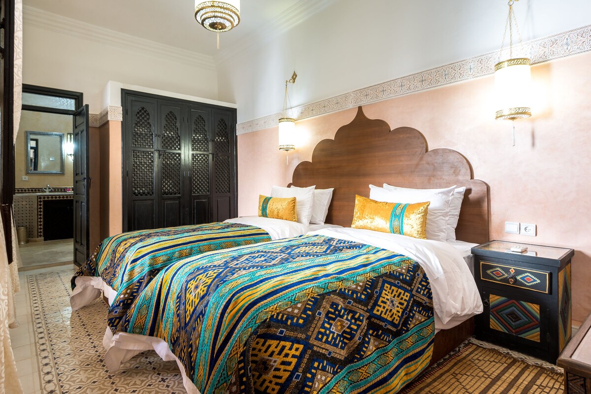 A hotel room with a large bed, a wooden headboard, patterned bedding, a nightstand, and a decorative wall panel.
