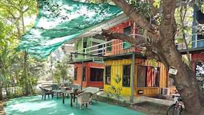 A colorful outdoor seating area with tables and chairs, surrounded by trees and a green tarp. - IORA Cottages (Alibag)