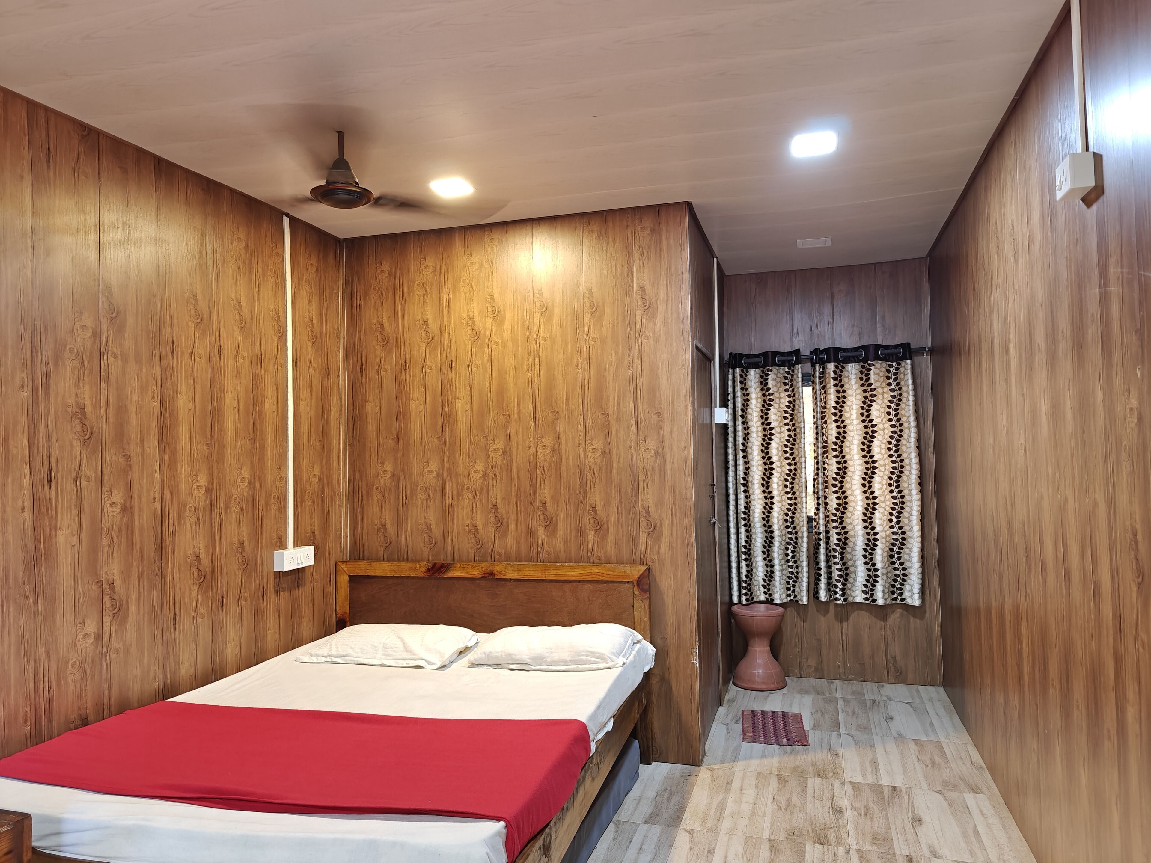Une chambre avec des murs en bois, un lit avec une literie blanche et rouge, un ventilateur de plafond et une fenêtre avec des rideaux à motifs.