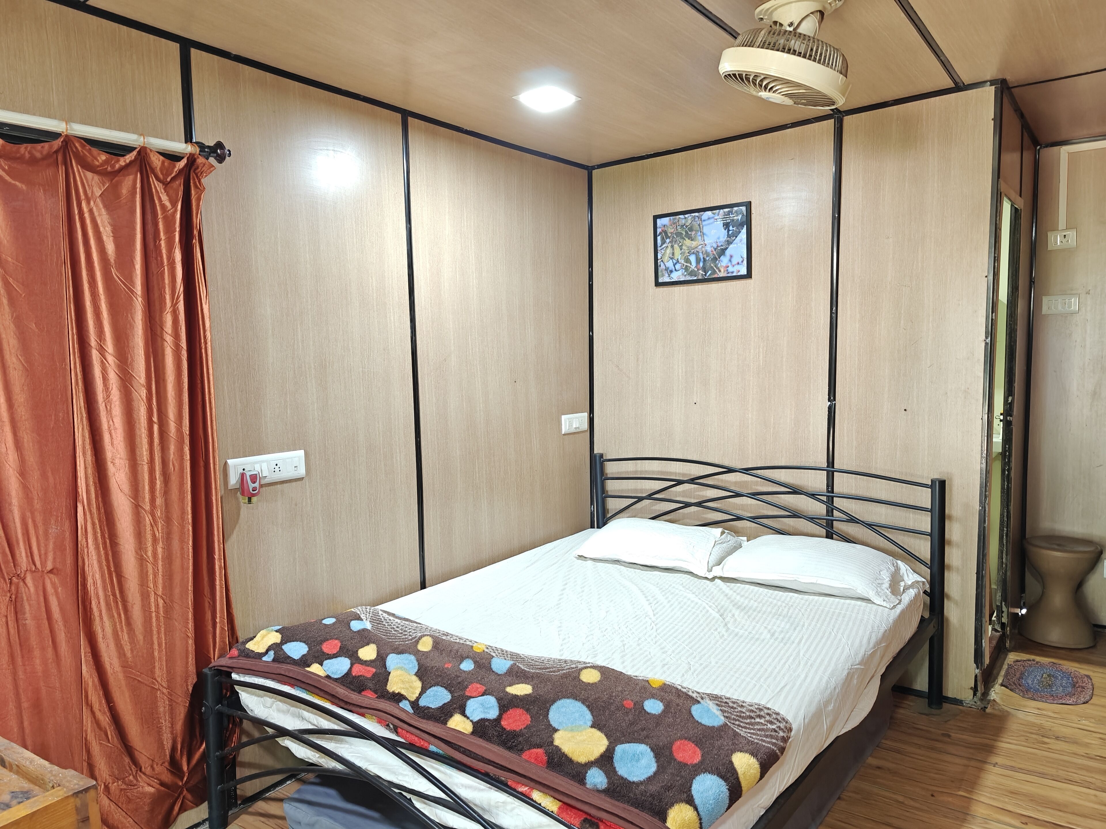 Un lit simple avec une couverture colorée, un ventilateur de plafond et un petit tableau accroché au mur.