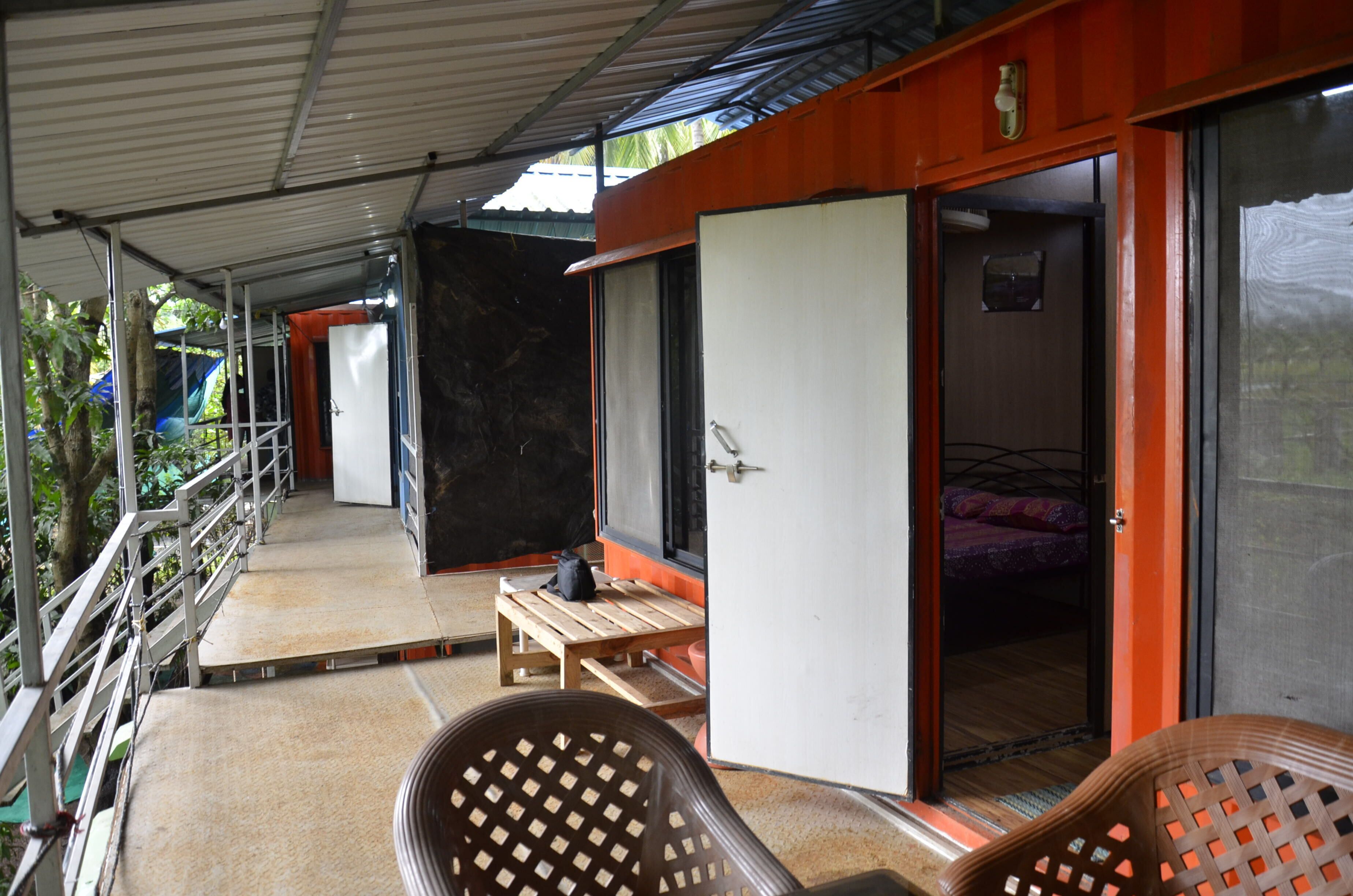 A covered outdoor area with a wooden bench, a chair, and a bed inside a room with an open door.