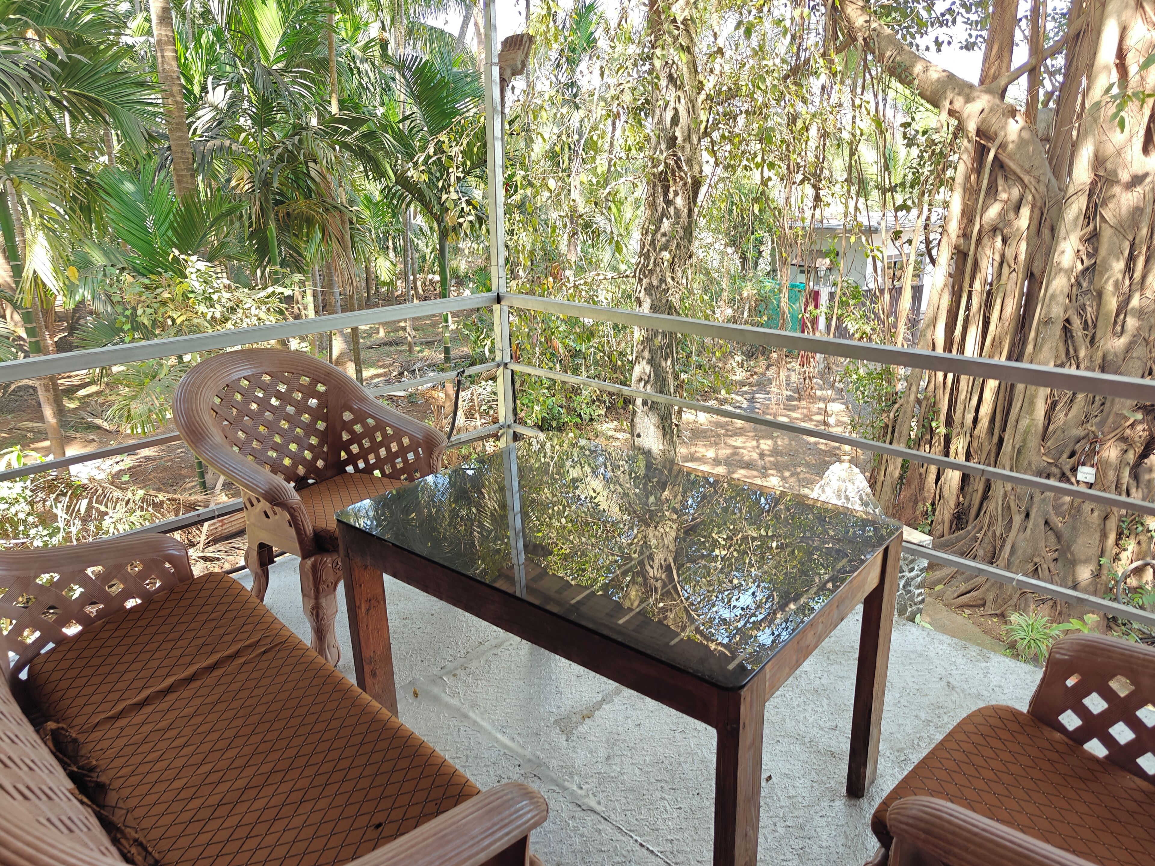 A balcony with a glass table and two wicker chairs, surrounded by lush greenery and trees.