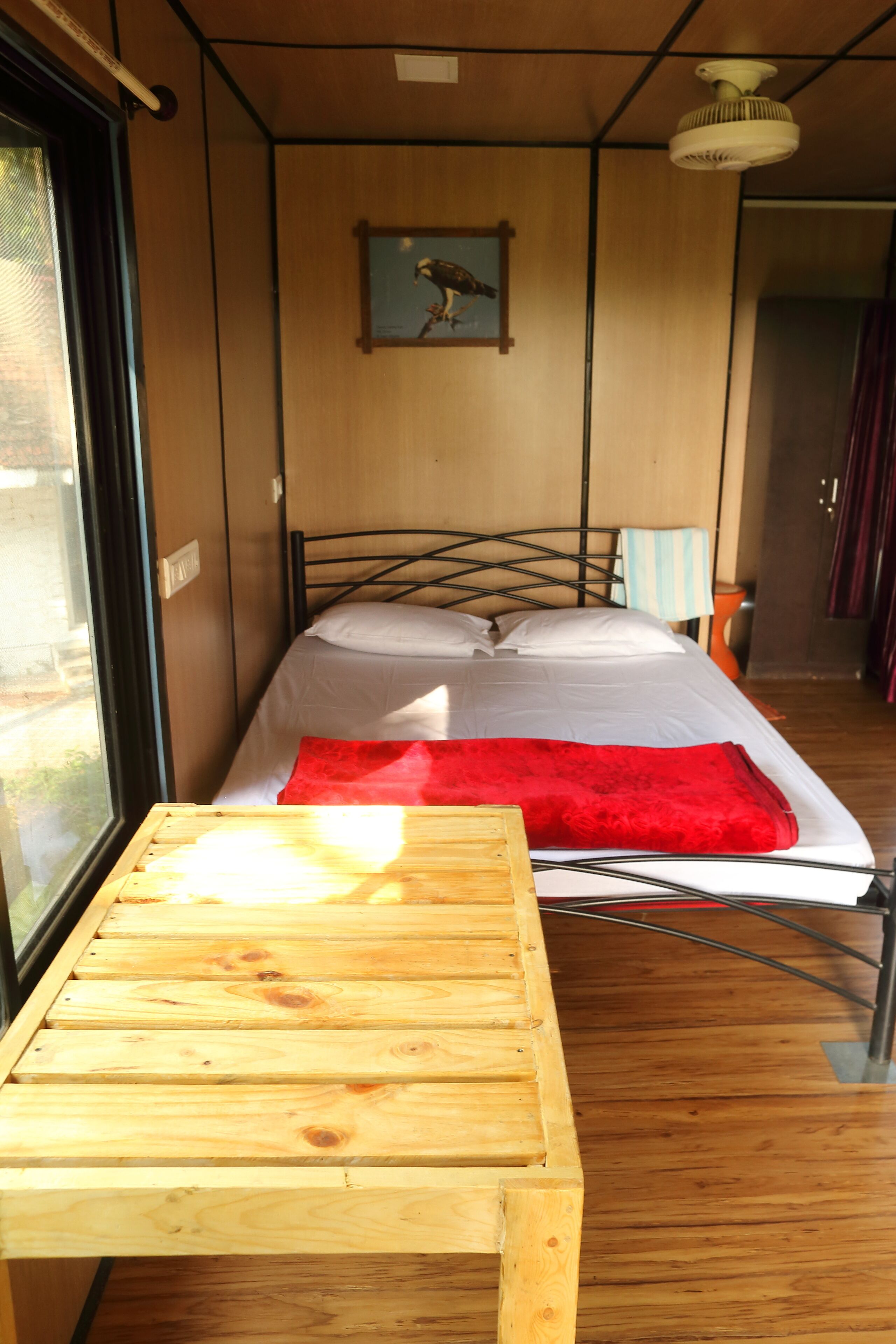 A small room with a bed, a wooden table, a window, and a ceiling fan.