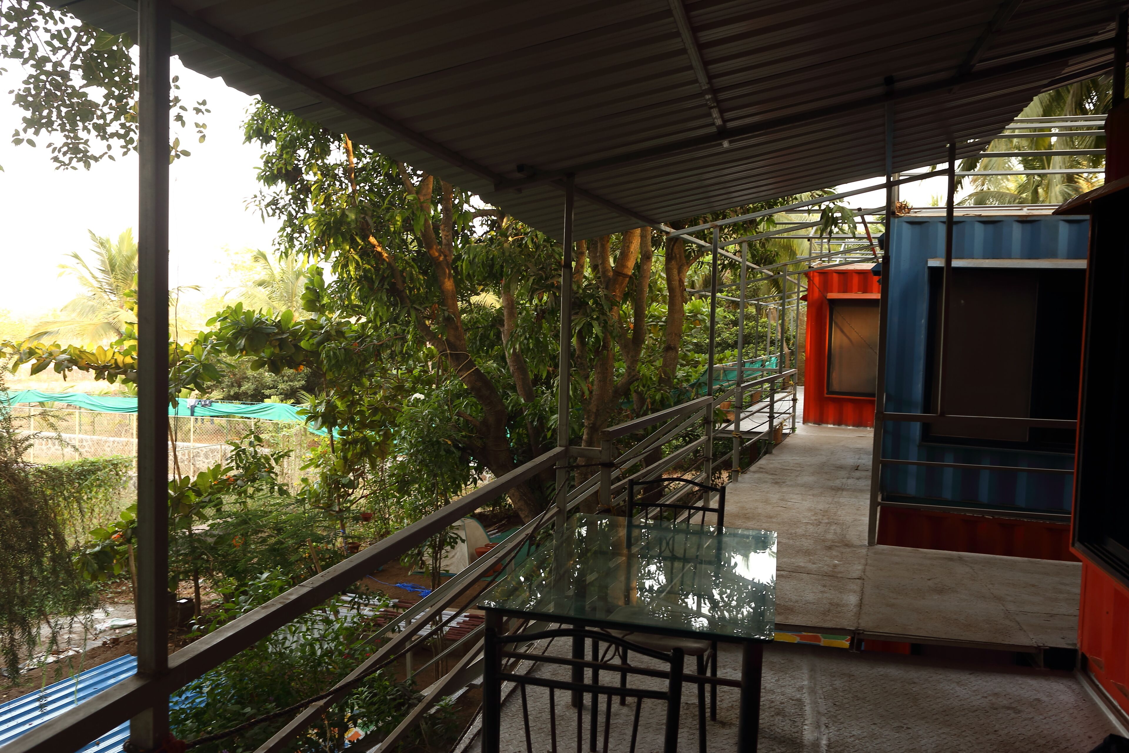A covered outdoor area with a glass table and chairs, surrounded by metal railings and trees.