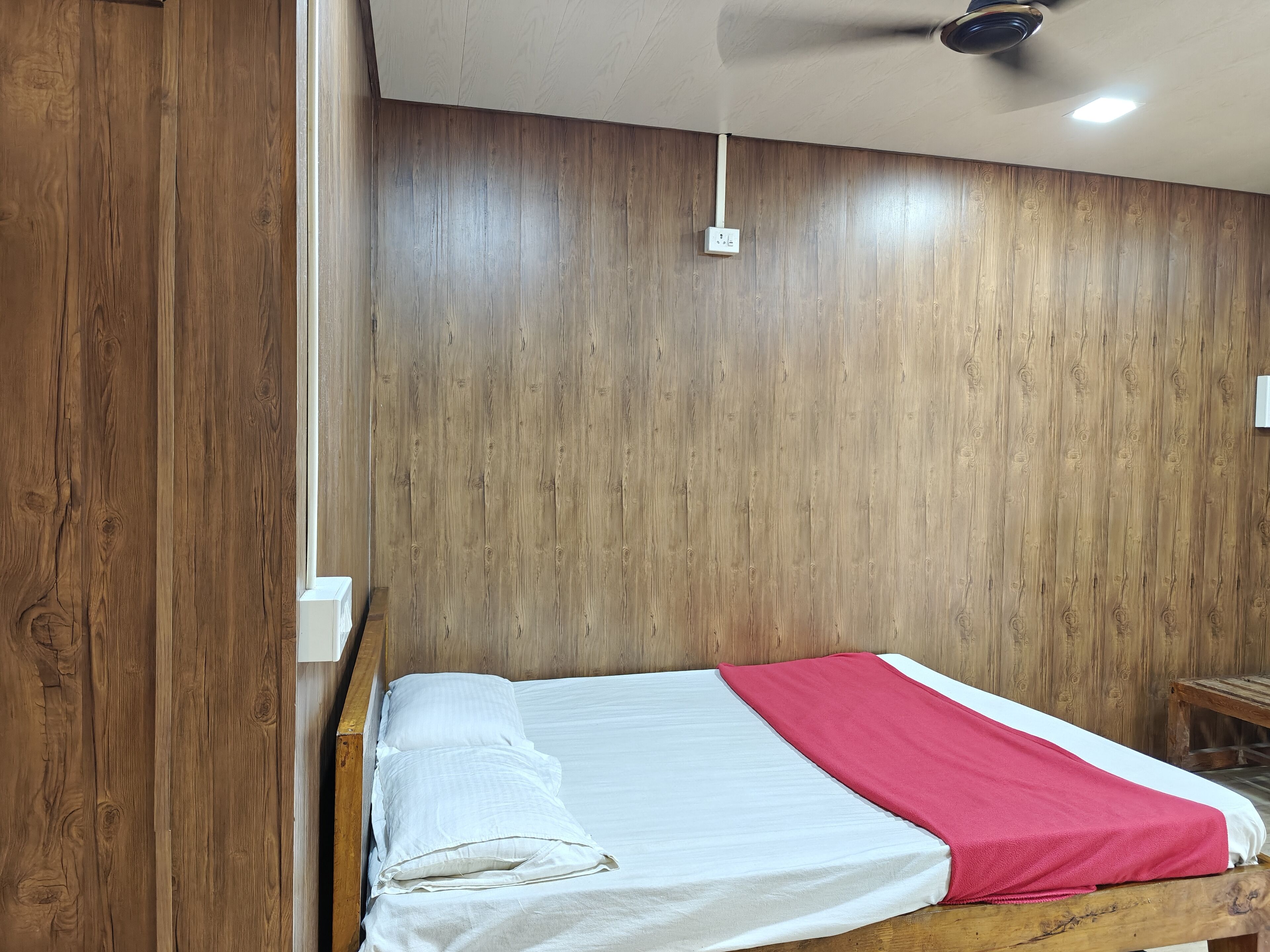 A single bed with a white and red bedspread in a room with wooden panel walls and a ceiling fan.