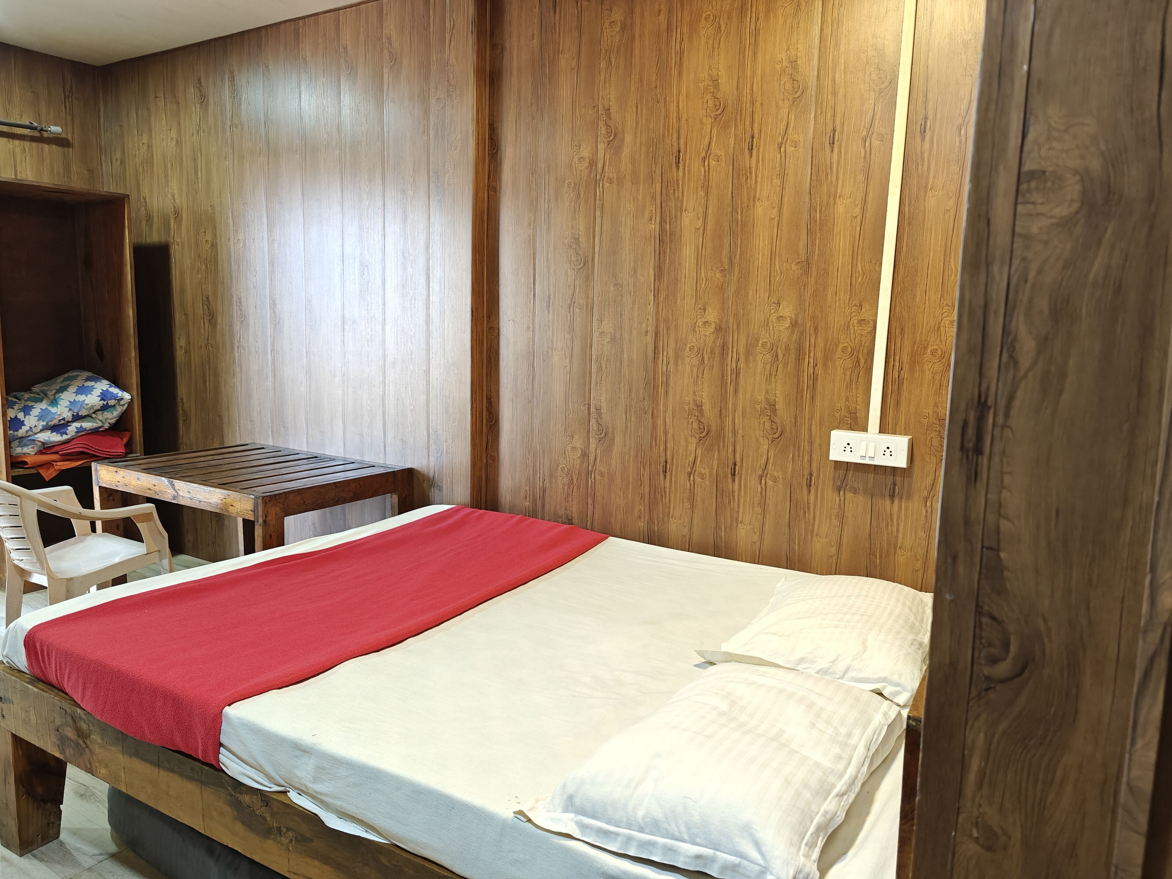Una cama individual con una manta roja, una mesa de madera y una silla en una habitación con paredes de madera y un armario.
