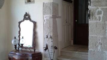 A hallway with a stone wall, a wooden dresser with a mirror, a vase with flowers, and a door at the end.