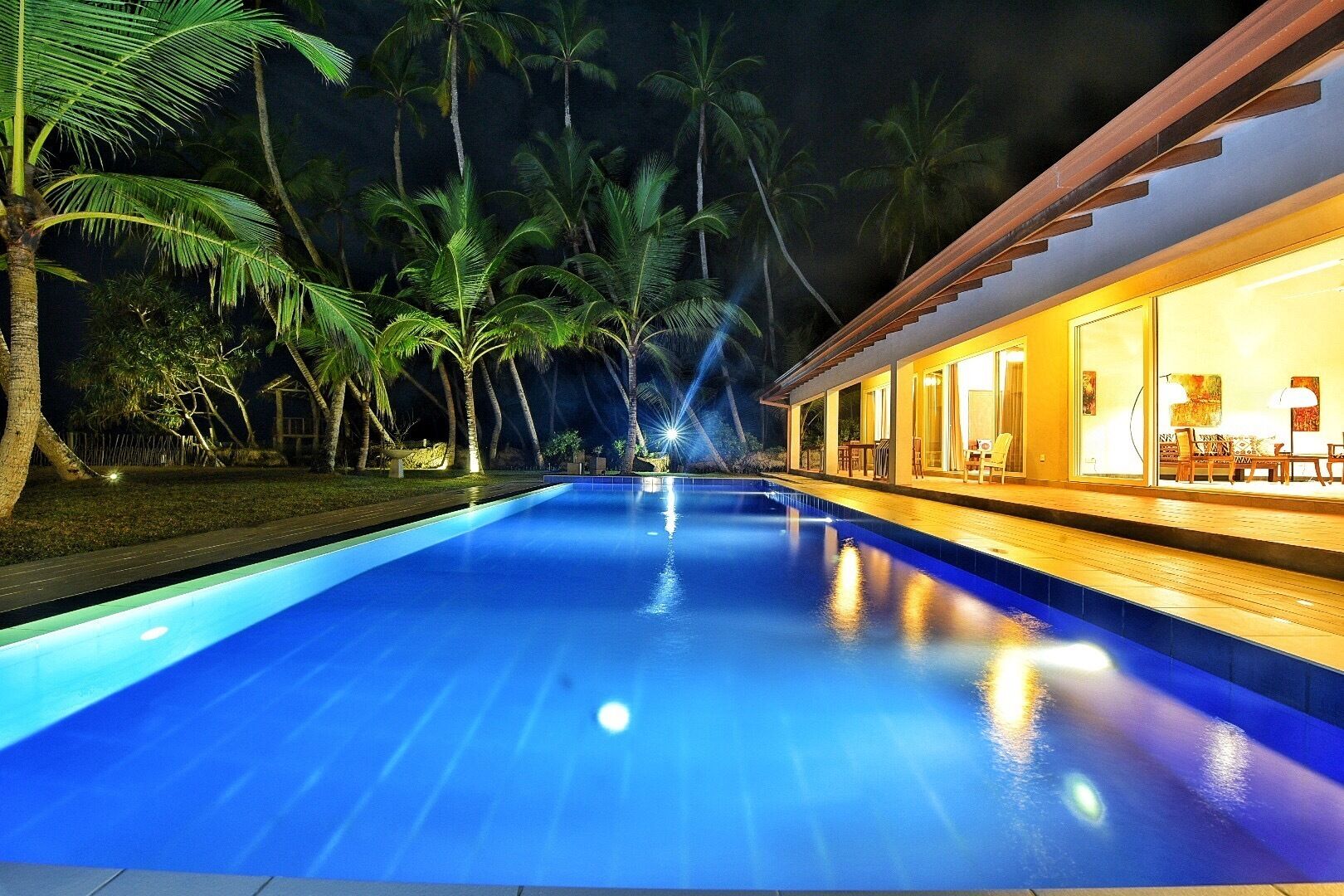 Una piscina nocturna con palmeras y un edificio con ventanas iluminadas.