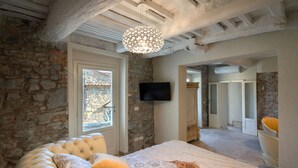 A bedroom with stone walls, a bed with a floral duvet, a television, and a chandelier. - Casa Collodi Castello (Collodi)