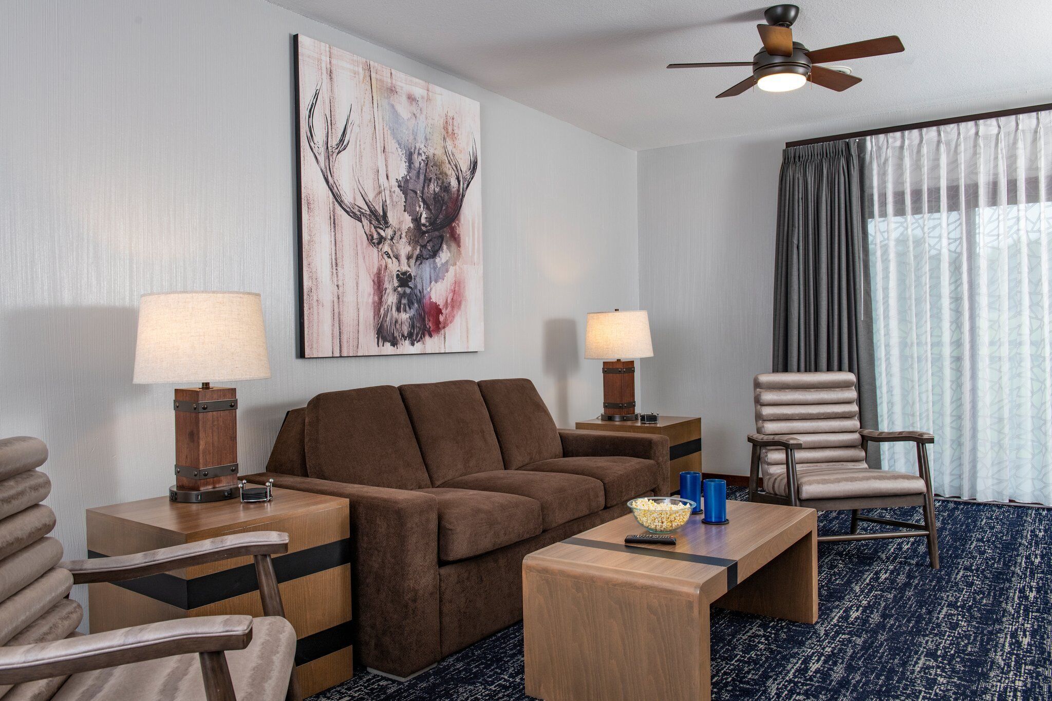 A hotel room with a brown sofa, two side tables with lamps, a wooden coffee table, a chair, and a ceiling fan.