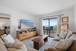 Una sala de estar con televisor grande, muebles de madera, balcón con vistas y comedor.
