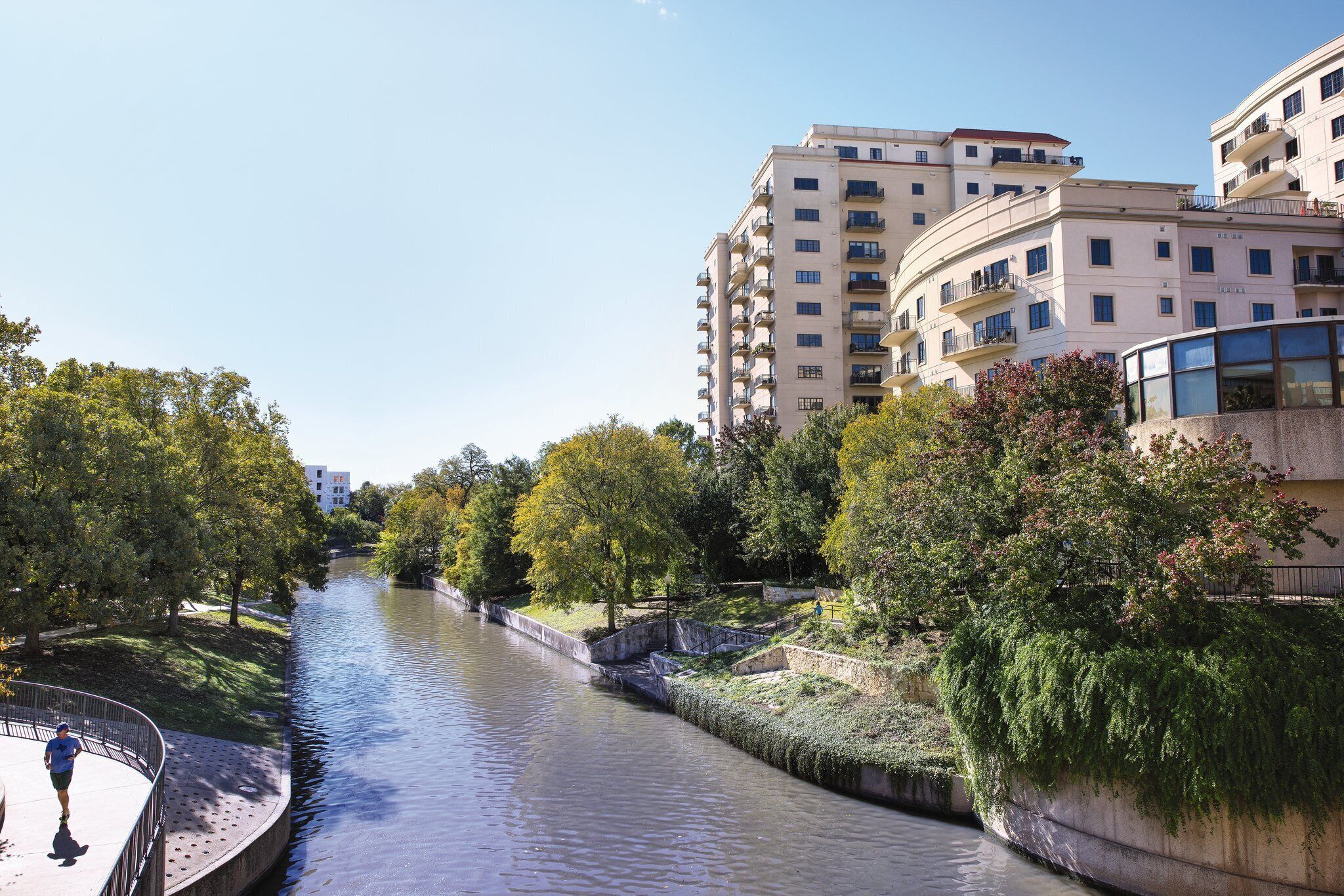 A riverside walkway with a bridge, surrounded by trees and modern buildings.