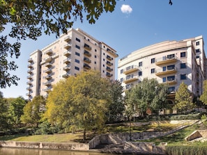 A multi-story residential building with balconies, surrounded by trees and a small water feature.