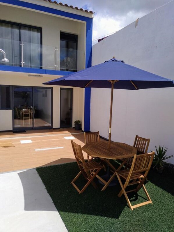 Um pátio com mesa e cadeiras de madeira, um guarda-sol azul e um pequeno vaso com planta.