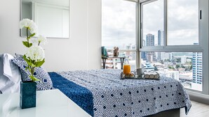 A bedroom with a large window, a bed with a patterned bedspread, a vase with flowers, and a city view.